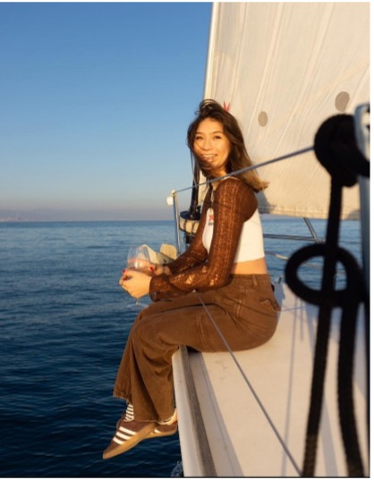 Person sitting on sailboat edge with ocean and horizon in background, holding a glass, smiling at sunset. Barcelona Sailing