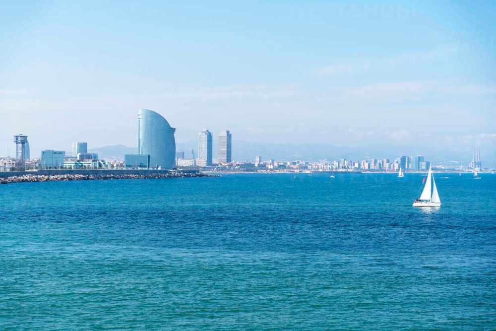 Coastal view of a city skyline with a sailboat on the blue ocean.