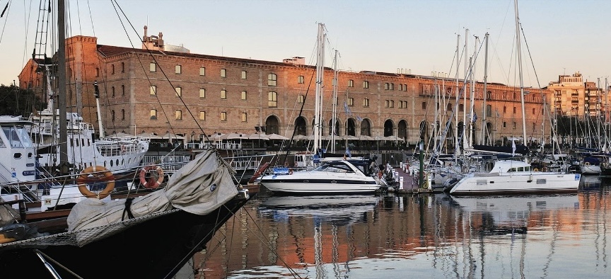 Boats moored in a calm marina with a large, red brick building in the background.