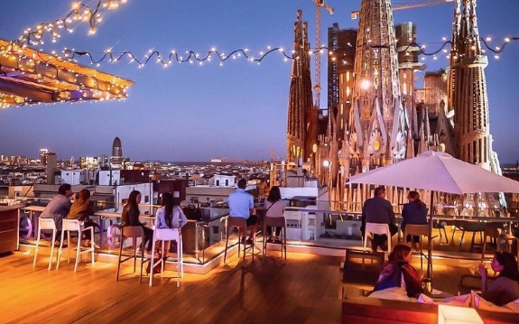 People dining on a rooftop terrace with cityscape and illuminated spires in the background at dusk.
