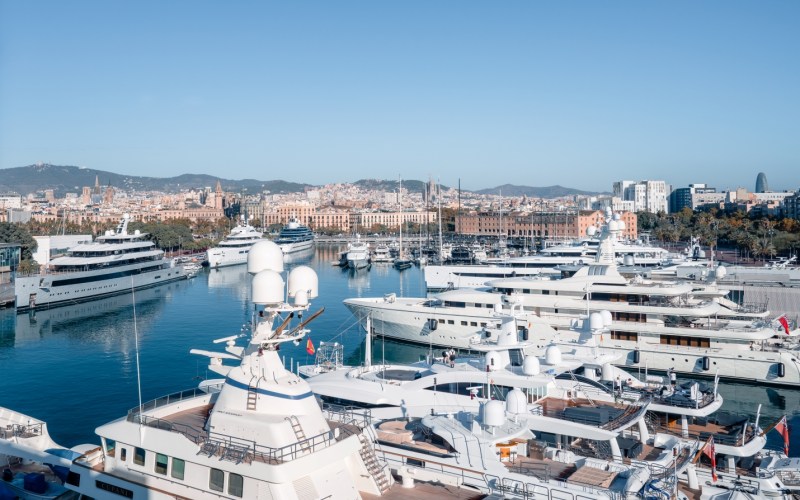 Yachts docked in a marina port vell with a Barcelona city skyline in the background under a clear blue sky.